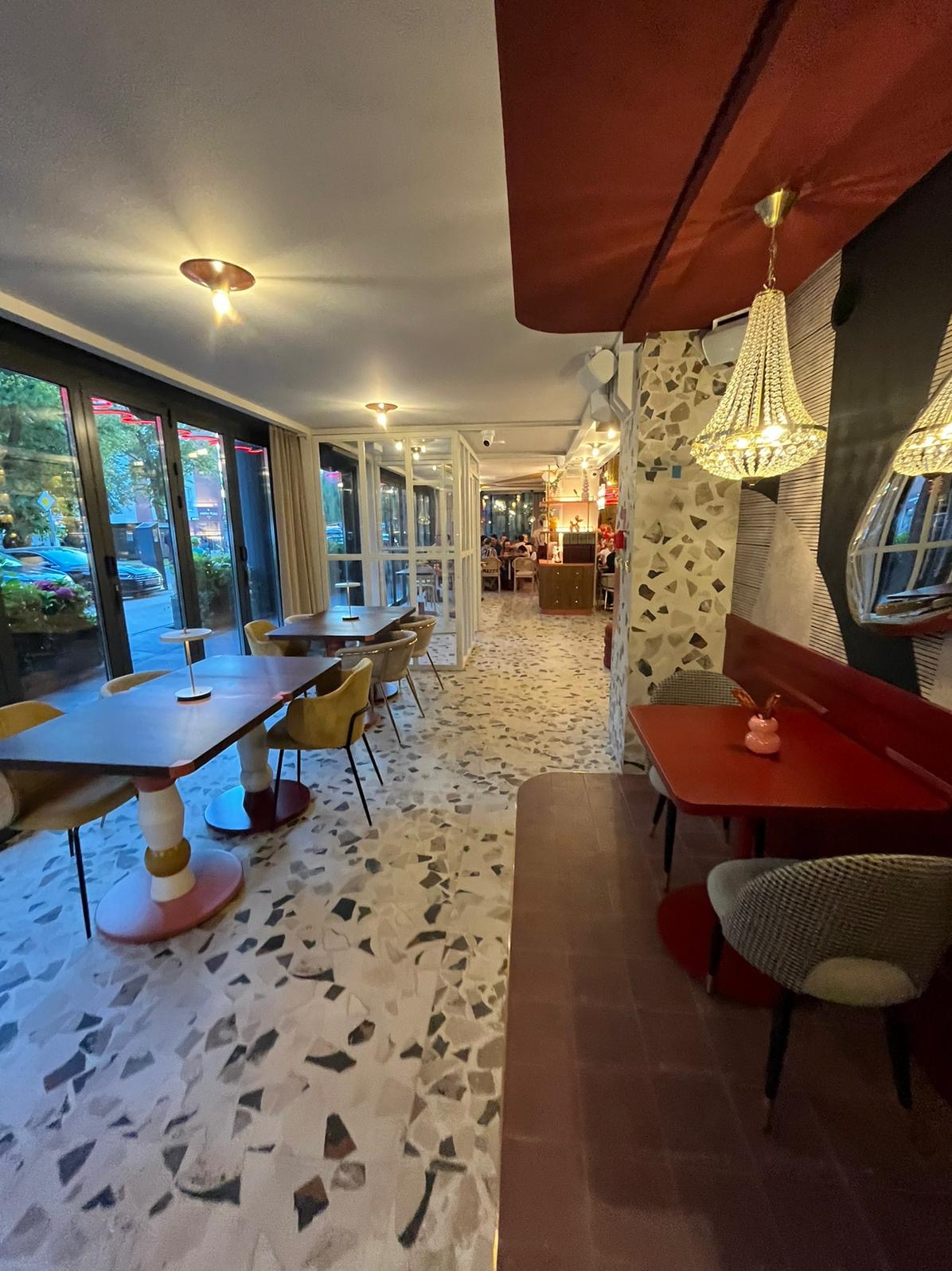 Wnętrze kawiarni z mozaikową podłogą, stolikami z żółtymi krzesłami i czerwonymi akcentami. Widoczne żyrandole i duże okna z widokiem na zieleń.