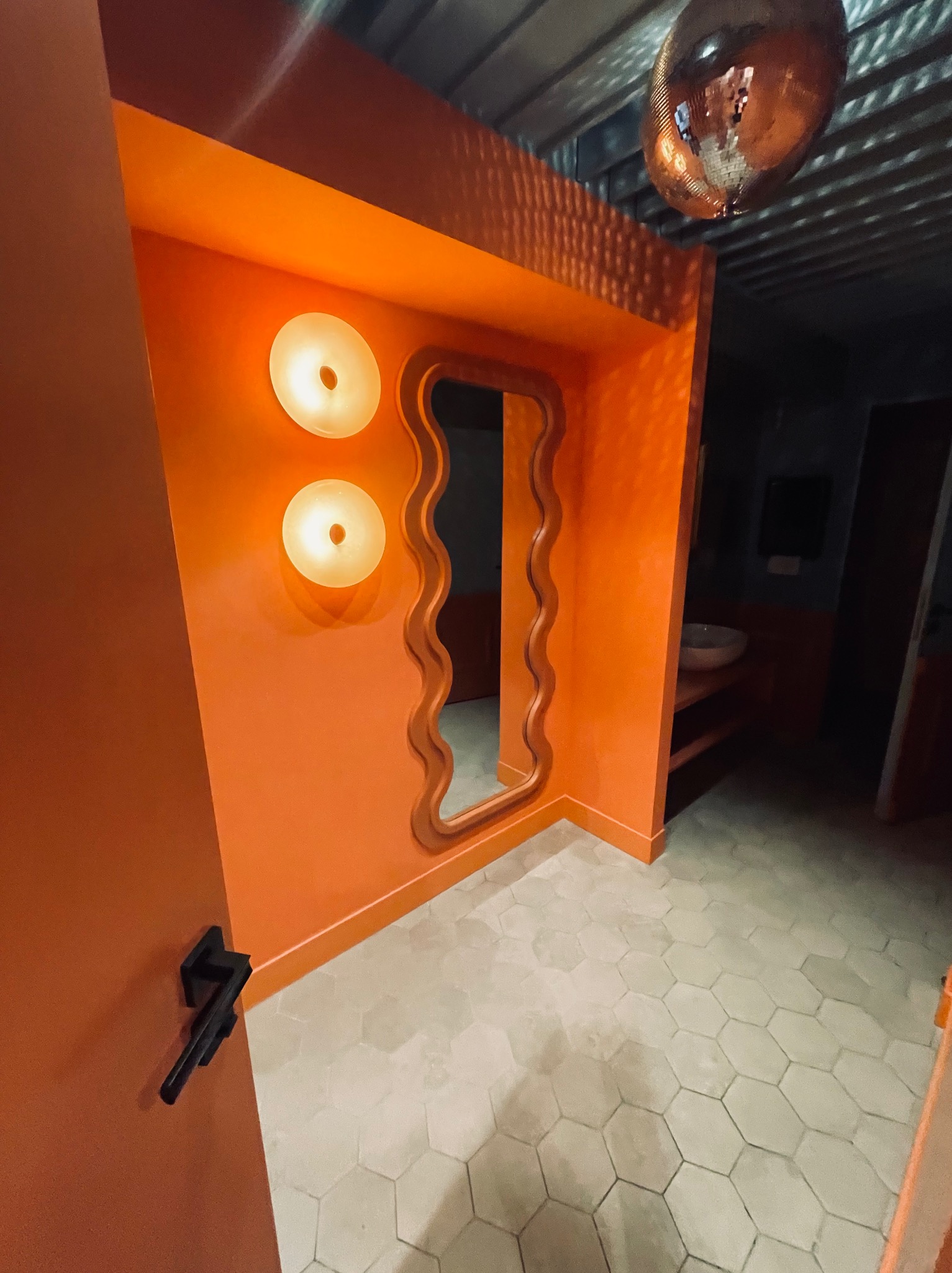 Pomarańczowa łazienka z lustrem o falistych krawędziach i heksagonalnymi płytkami podłogowymi. Dwie okrągłe lampy na ścianie, kula dyskotekowa na suficie.