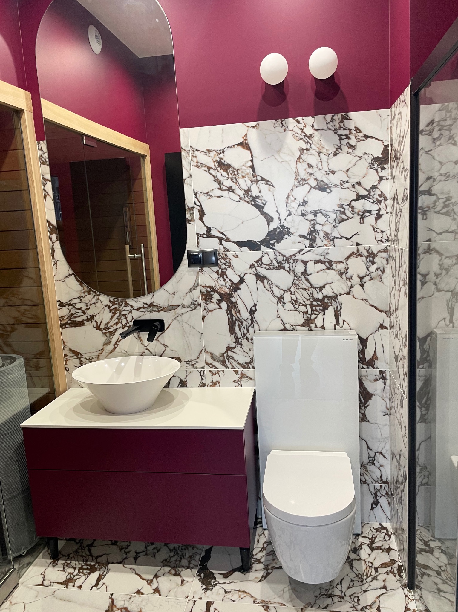 Nowoczesna łazienka z marmurowymi płytkami, burgundowymi ścianami i szafką pod umywalkę. Uwagę przykuwa owalne lustro z drewnianą ramą i minimalistyczna armatura.