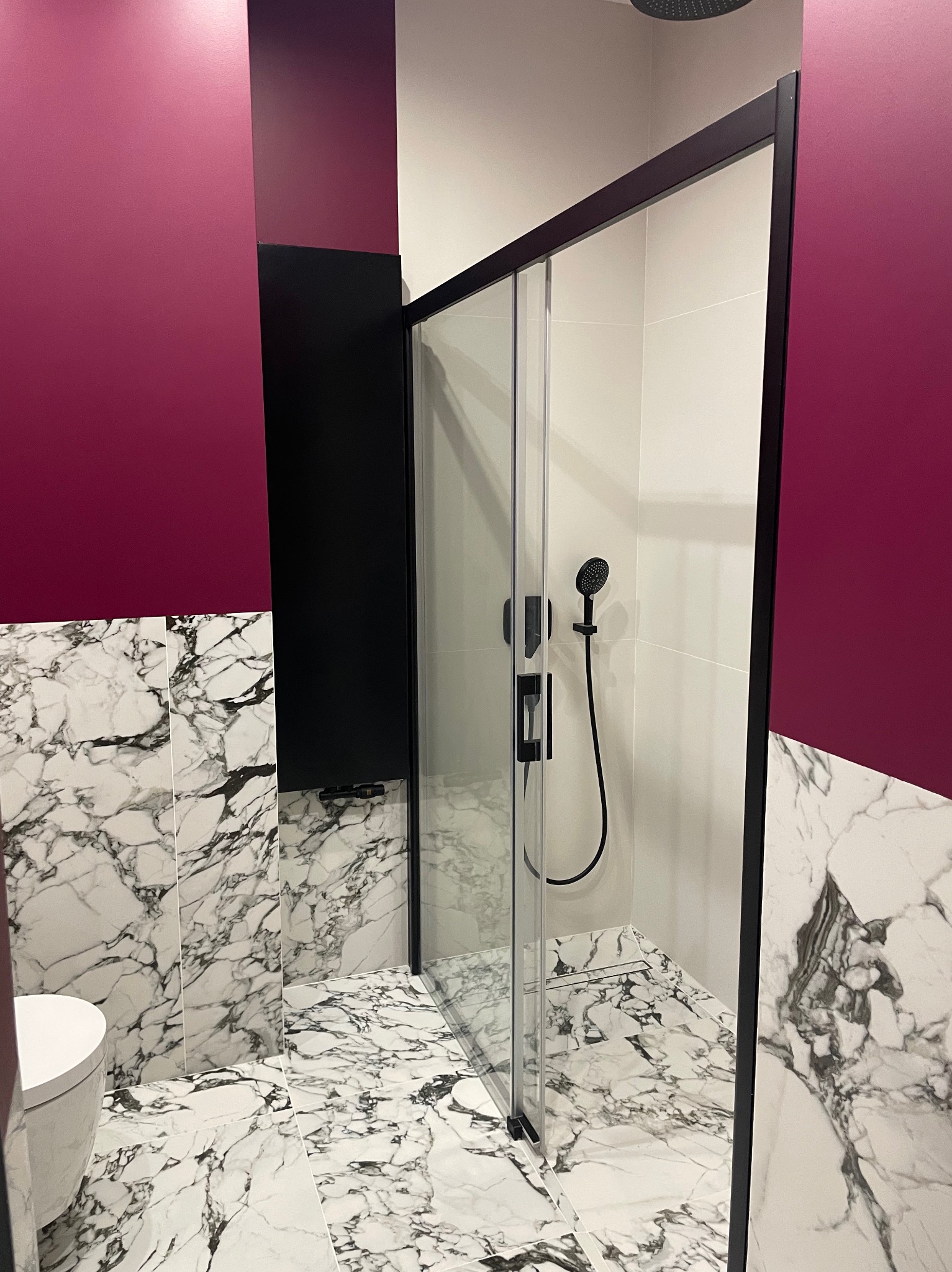Nowoczesna łazienka z kabiną prysznicową typu walk-in, marmurowymi płytkami i bordową ścianą. Czarna armatura i minimalistyczny design.