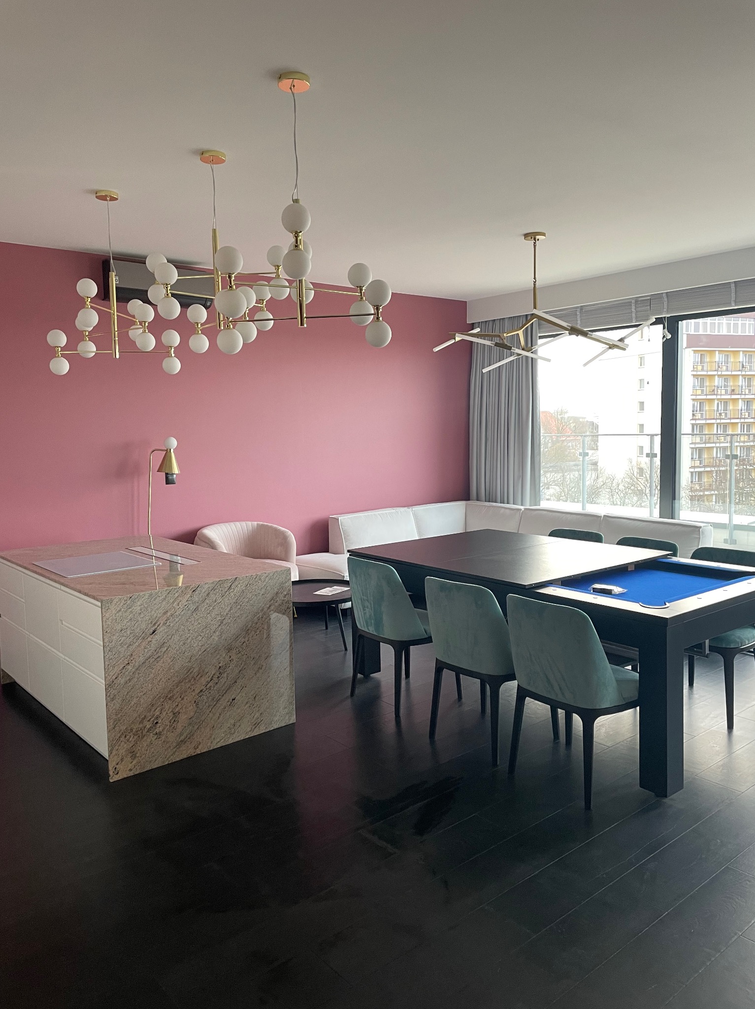 Nowoczesne wnętrze: różowa ściana, designerskie lampy, stół bilardowy z błękitnym suknem, minimalistyczna kuchnia z kamiennym blatem i czarną podłogą.