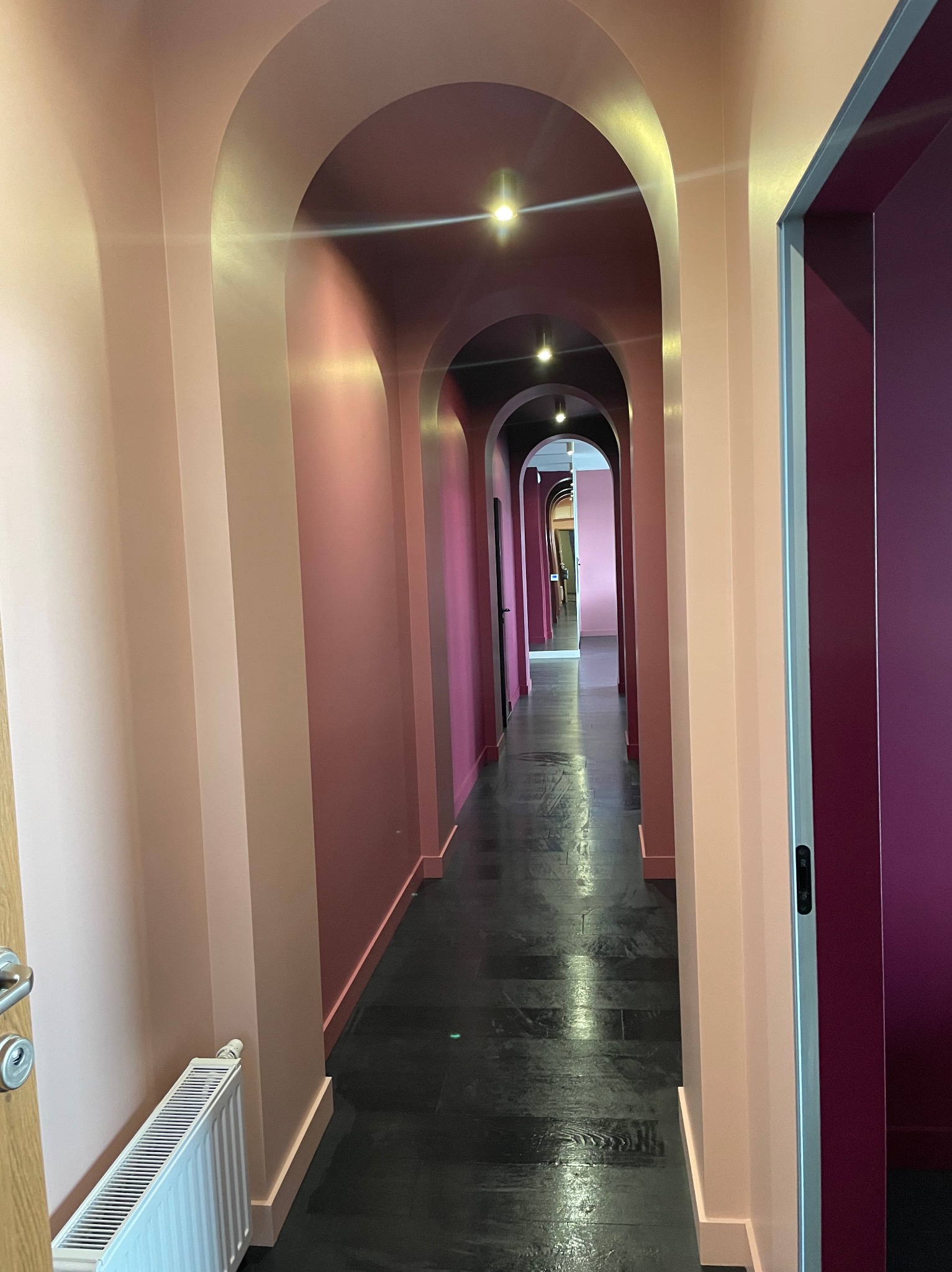 Aranżacja korytarza z łukami w odcieniach różu i burgundu, czarna podłoga z połyskiem. Grzejnik na ścianie po lewej stronie kadru.