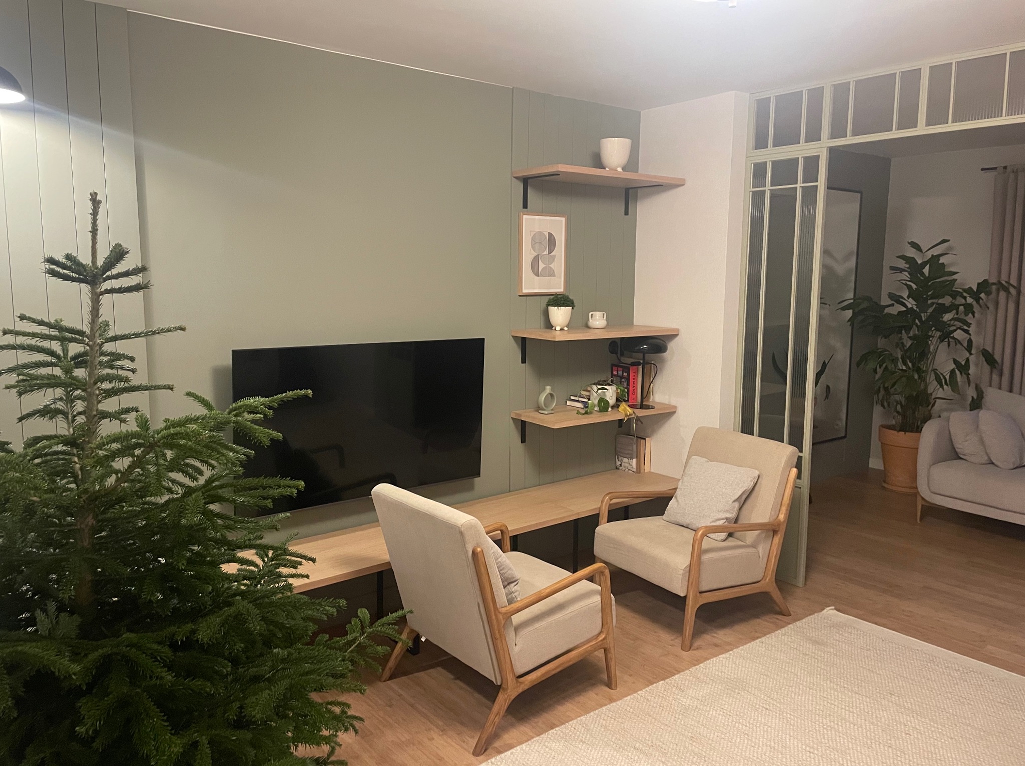 Nowoczesny salon z zieloną ścianą, drewnianymi półkami i telewizorem. Dwa beżowe fotele i choinka tworzą przytulną atmosferę. Widoczne detale wykończenia wnętrza.