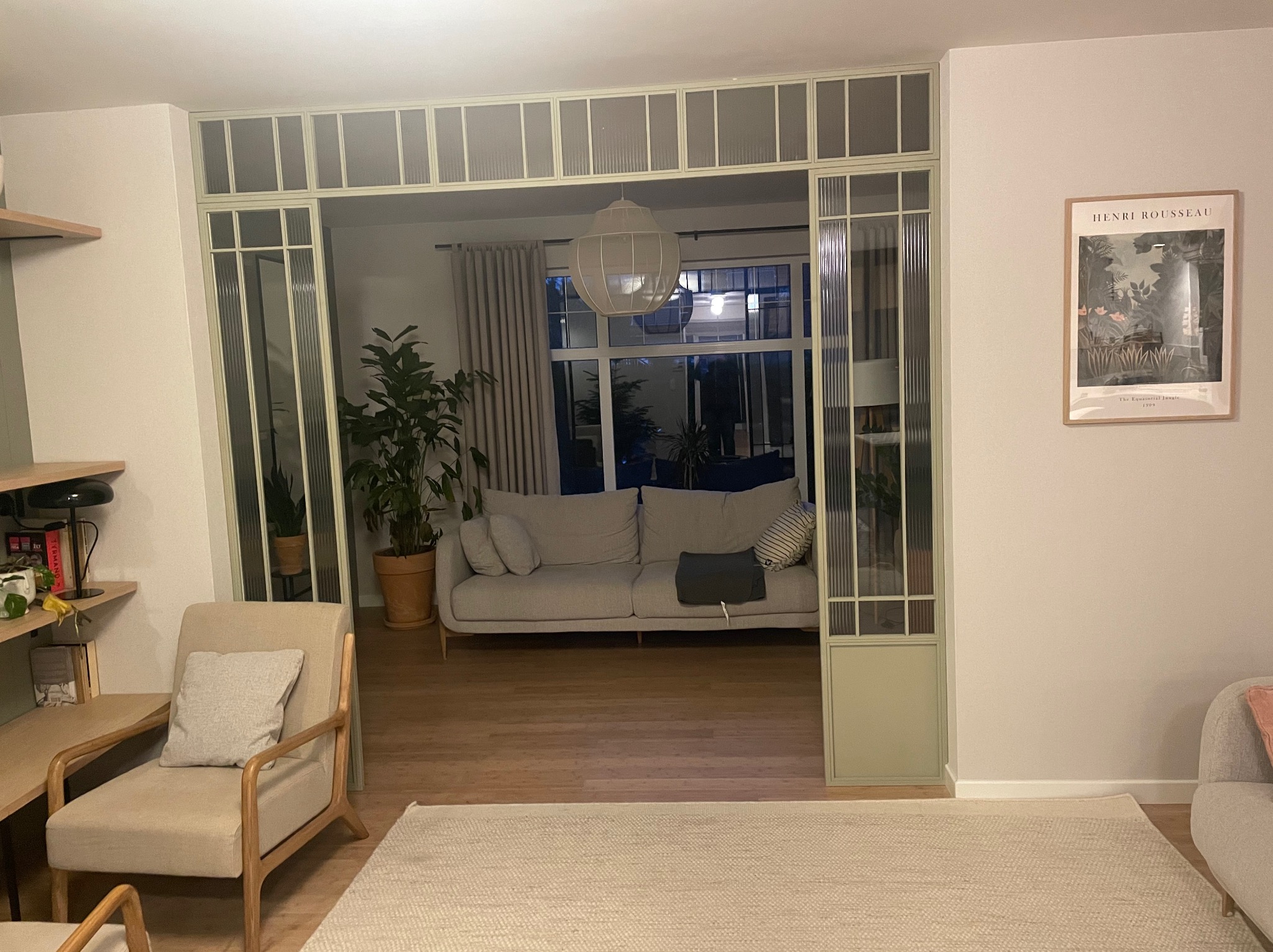 Stylowe wykończenie wnętrza: salon z przeszklonymi drzwiami w odcieniach zieleni, beżowy dywan, sofa i fotel. Na ścianie plakat Henri Rousseau.