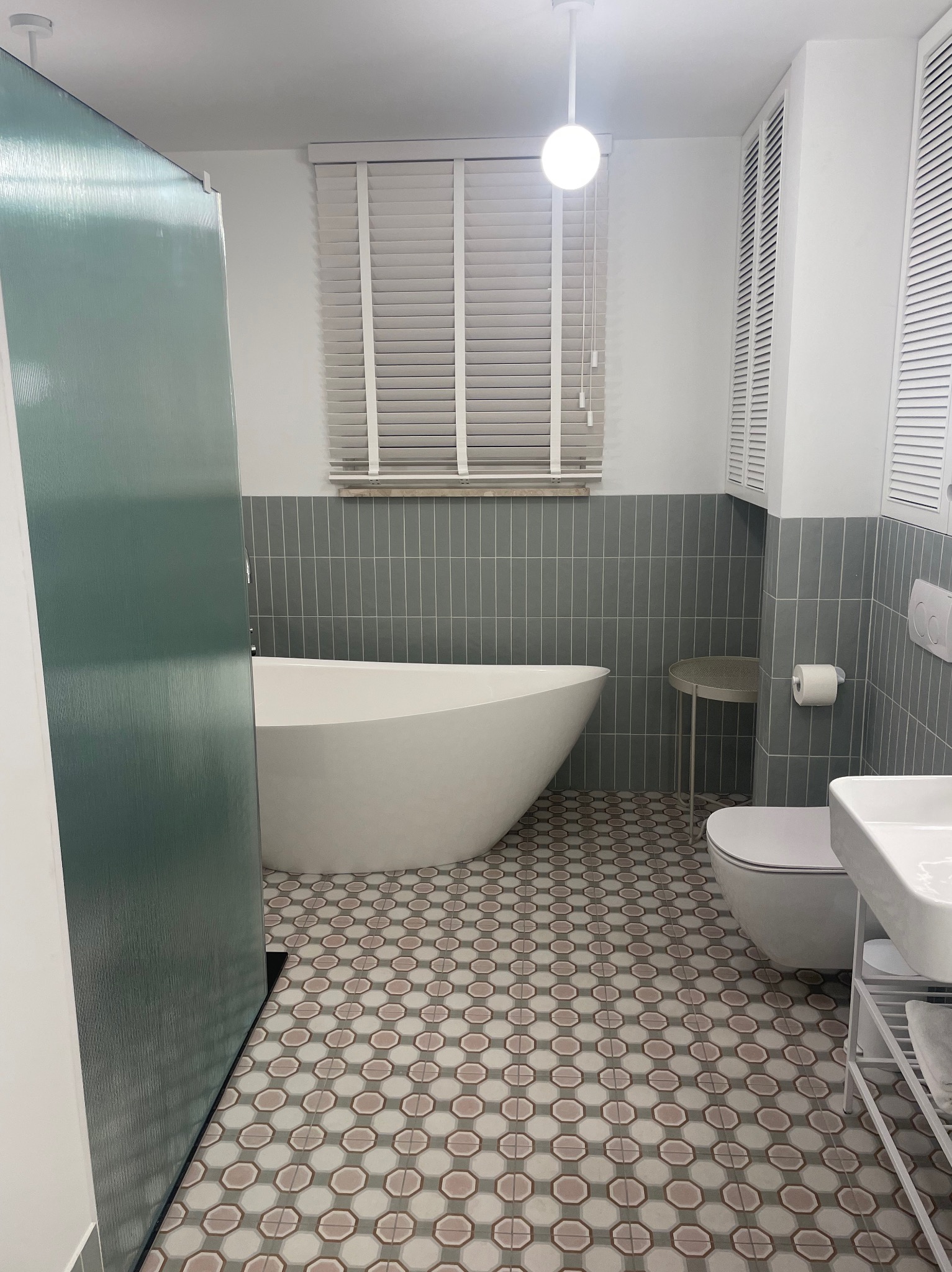 Nowoczesna łazienka z wolnostojącą wanną, szarą glazurą i wzorzystą podłogą. Widoczna kabina prysznicowa z zielonkawego szkła i białe żaluzje.