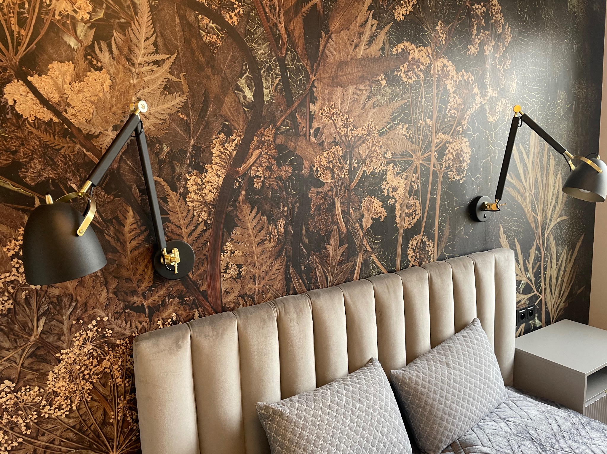 Wzorzysta tapeta w motywy roślinne za łóżkiem z zagłówkiem. Dwie czarne lampki nocne z elementami złota. Widoczny fragment szafki nocnej i pościeli.