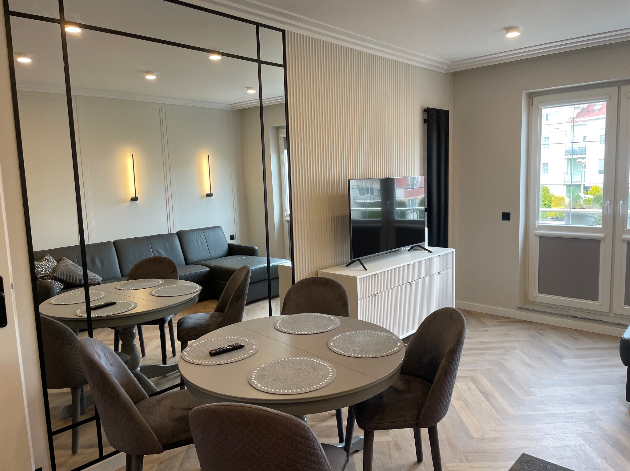 Stylowe wnętrze: salon z okrągłym stołem, krzesłami z aksamitu, telewizorem i dekoracyjną ścianą. Duże lustro oddziela przestrzeń. Parkiet ułożony w jodełkę.