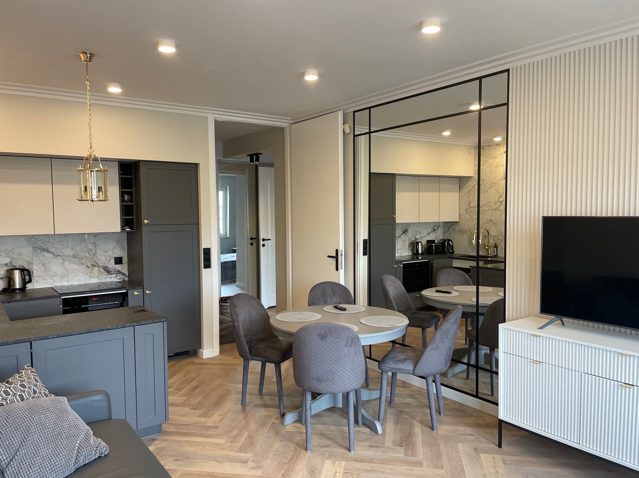 Stylowe wnętrze: szara kuchnia z marmurowym backsplash, okrągły stół z krzesłami, telewizor na białej komodzie z frezowanymi frontami i parkiet w jodełkę.