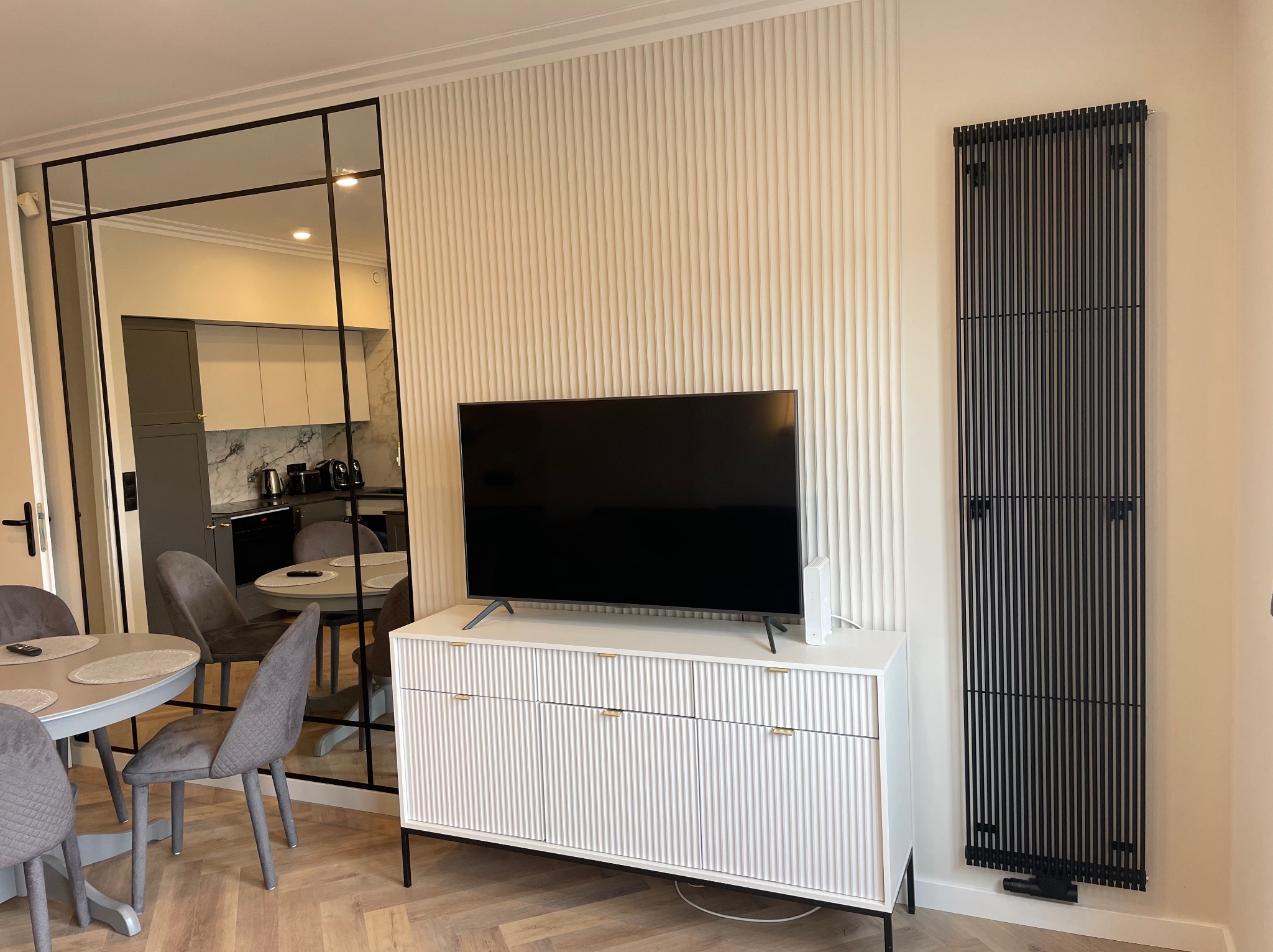 Nowoczesny salon po remoncie: ściana z lamelami, czarny grzejnik panelowy i biała komoda pod telewizorem. W lustrze widać jadalnię z kuchnią w tle.