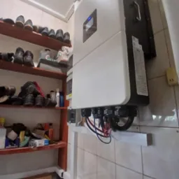 Biały inwerter fotowoltaiczny zamontowany na ścianie z kafelkami, widoczne kable. Obok półka z obuwiem i różnymi przedmiotami. Ujęcie z dołu, z bliska.