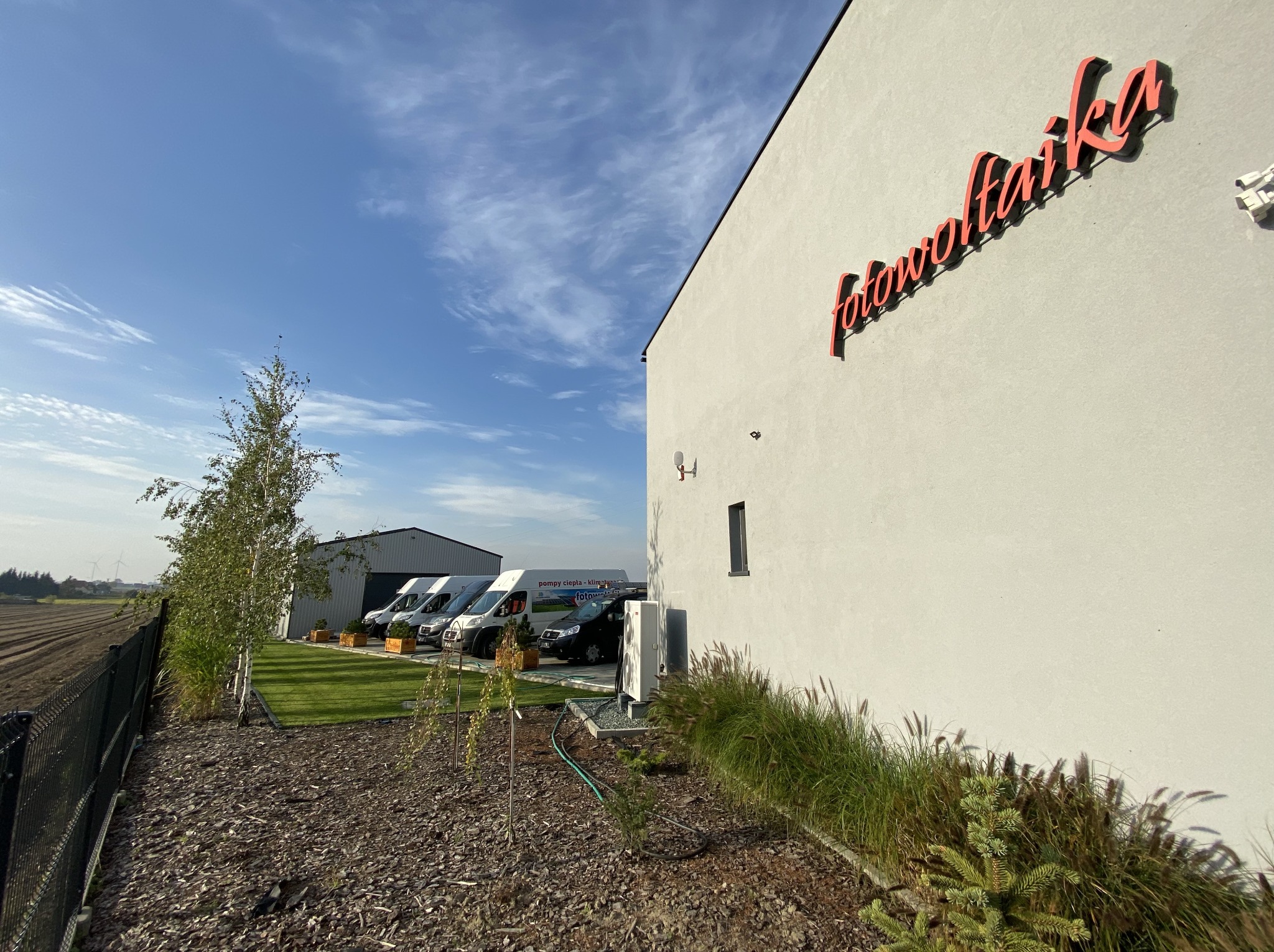 Budynek firmy z napisem 'fotowoltaika' na ścianie, kilka samochodów dostawczych zaparkowanych z boku, w tle pole i turbiny wiatrowe.