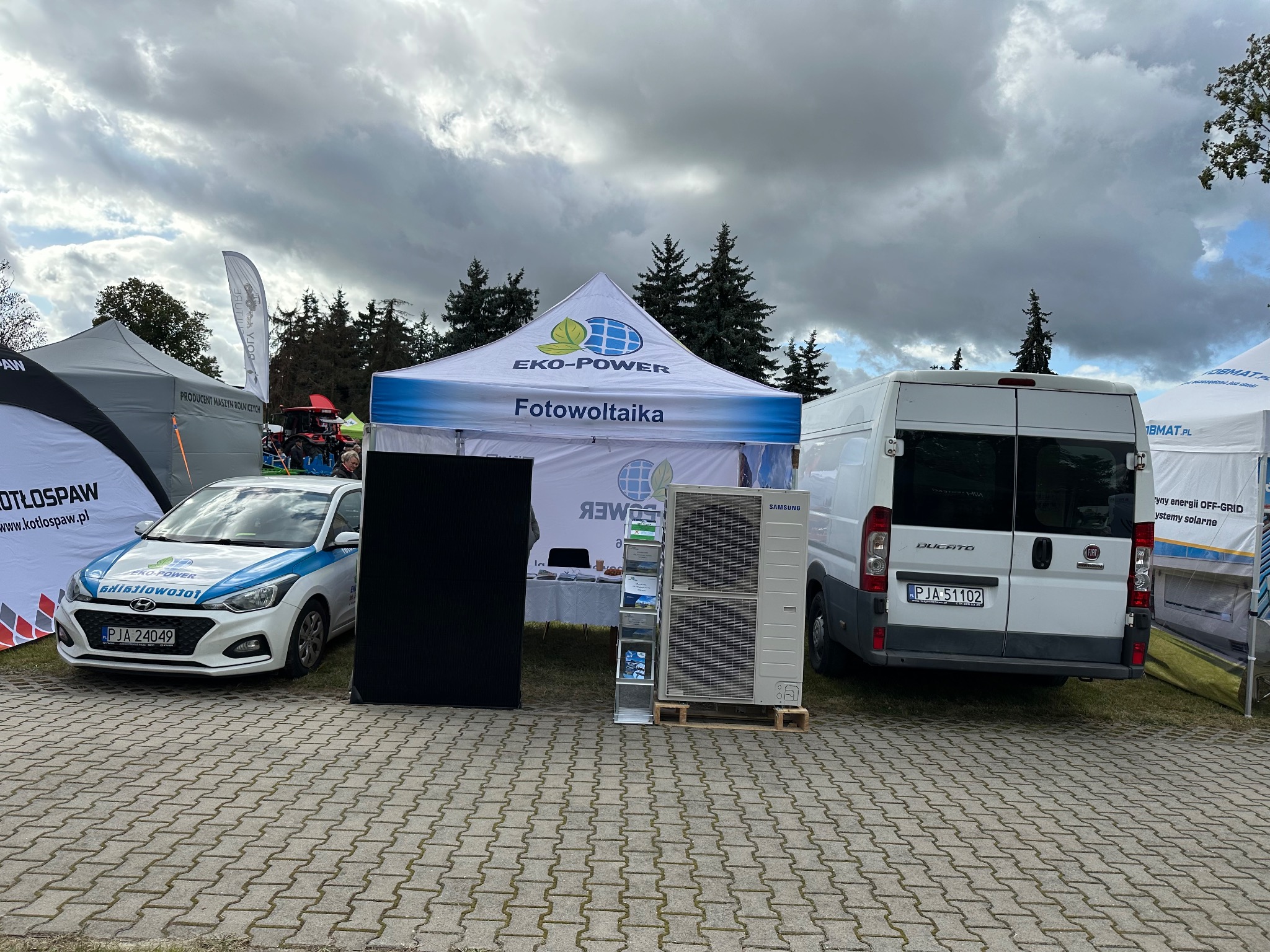 Ekspozycja firmy Eko-Power na zewnątrz: namiot z napisem Fotowoltaika, auto z logo, klimatyzator Samsung i bus Ducato na kostce brukowej, pochmurne niebo.