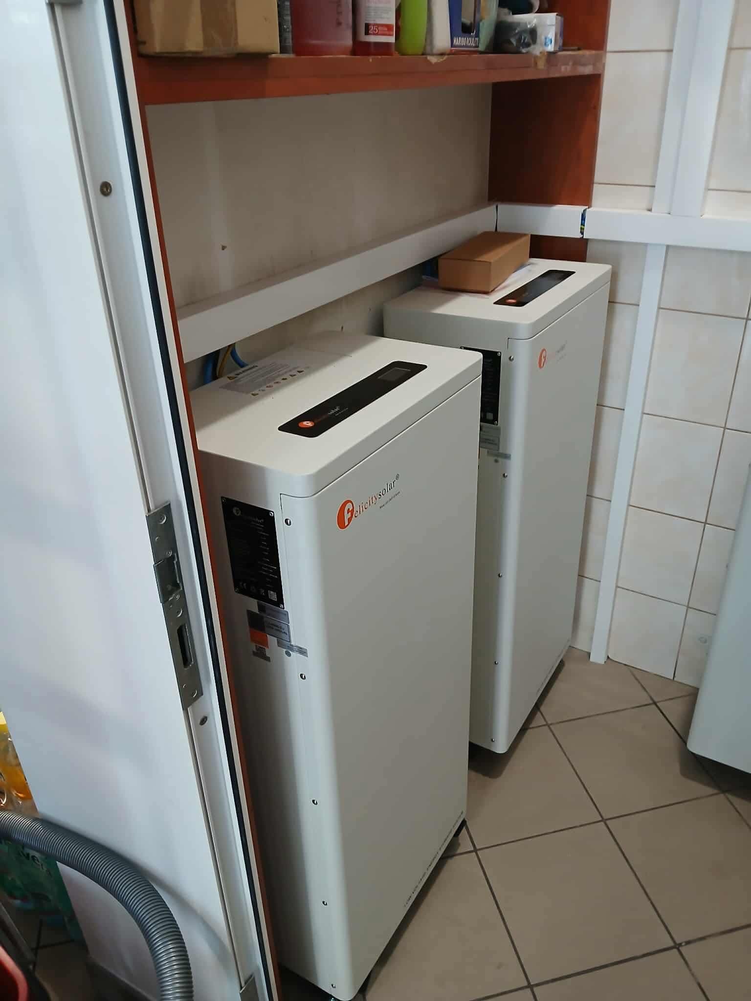 Dwa białe magazyny energii Felicitysolar ustawione obok siebie w pomieszczeniu z beżową podłogą i półkami. Widoczny fragment szafki i rury od odkurzacza.