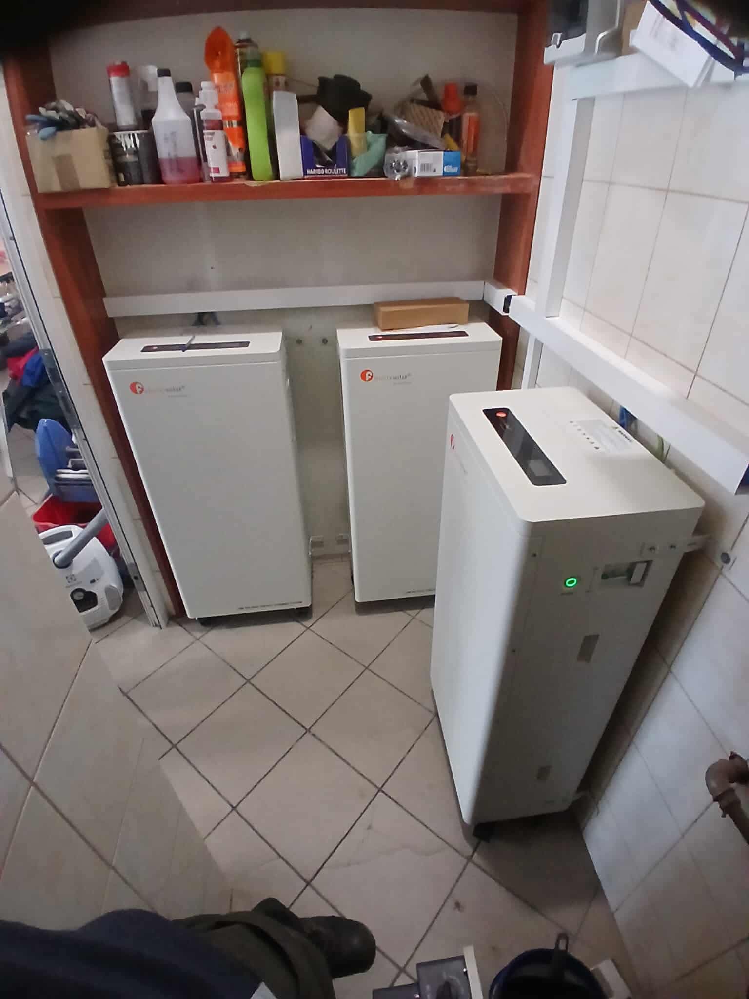 Trzy białe magazyny energii w pomieszczeniu z półką, widoczne przewody i rury. Jeden z magazynów z włączonym zielonym wskaźnikiem.