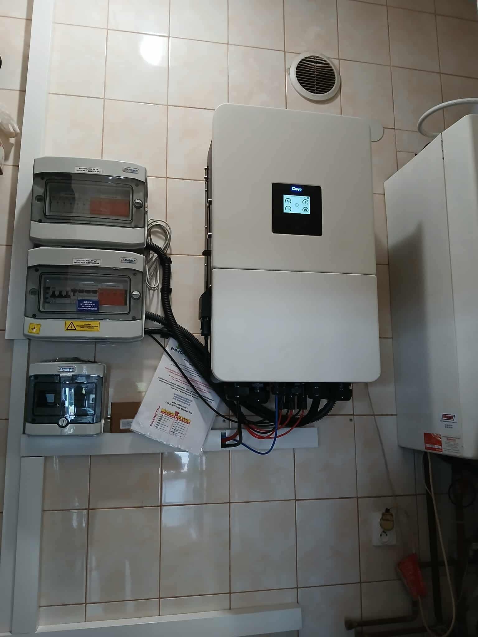Wnętrze z zamontowanym inwerterem fotowoltaicznym marki Deye, trzema rozdzielnicami elektrycznymi i fragmentem bojlera. Widoczne kable i dokumentacja.