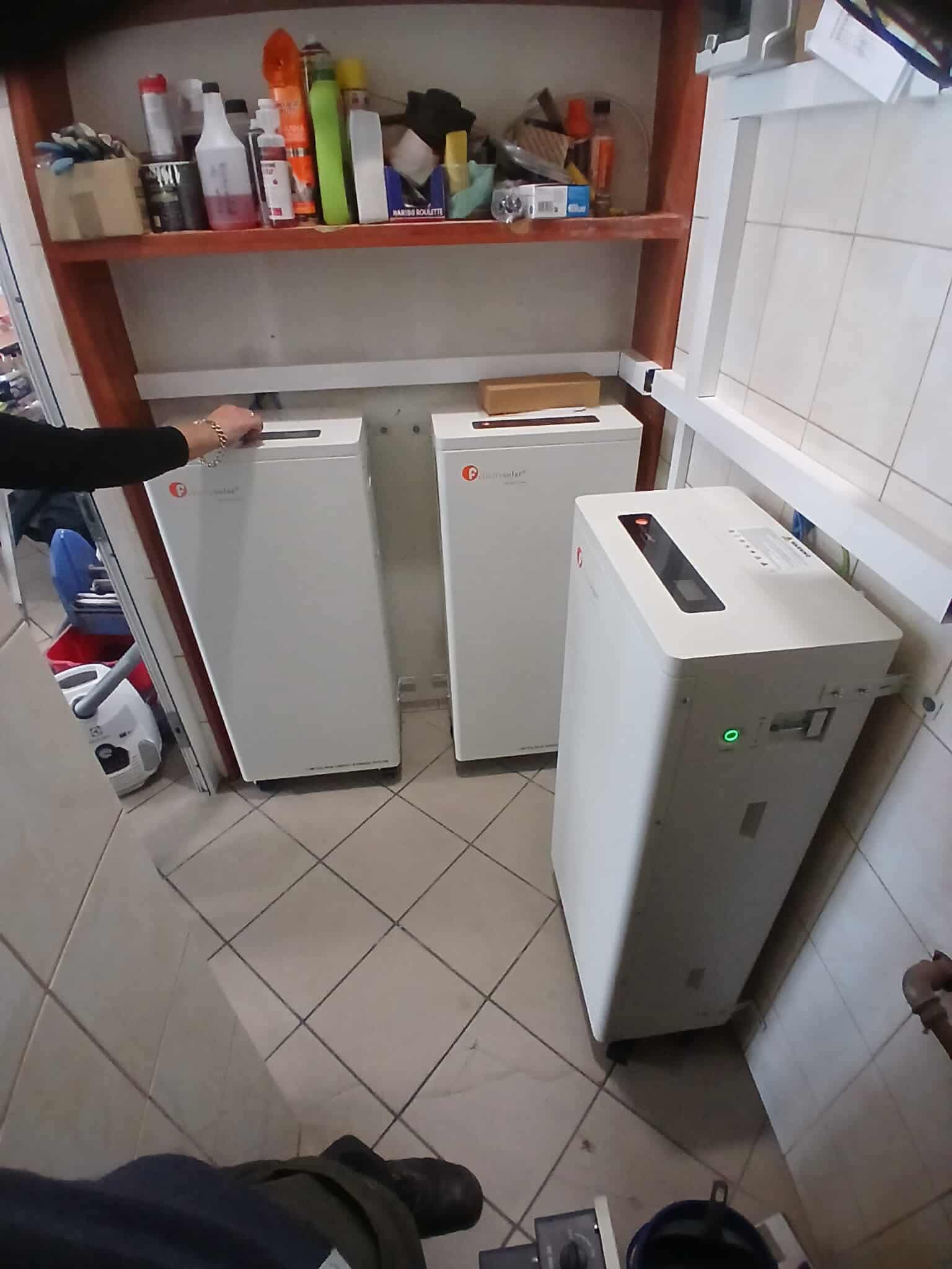 Trzy białe magazyny energii ustawione w pomieszczeniu z kafelkami. Ręka z bransoletką dotyka górnej części jednego z urządzeń. Widoczne półki z różnymi przedmiotami.