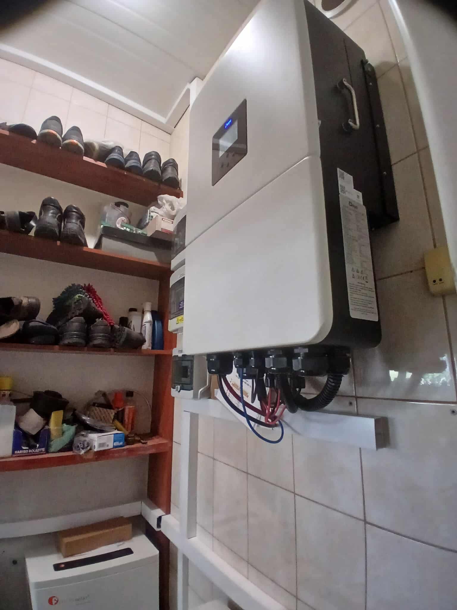 Biały inwerter fotowoltaiczny zamontowany na ścianie z kafelkami, widoczne kable. Obok półka z obuwiem i różnymi przedmiotami. Ujęcie z dołu, z bliska.