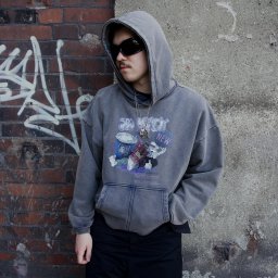 Dwa De Studio - Mężczyzna w bluzie z kapturem i nadrukiem DTF, efekt sprania. Tło: ceglana ściana z graffiti i betonowa ściana. Uwagę zwracają okulary przeciwsłoneczne i łańcuszek.