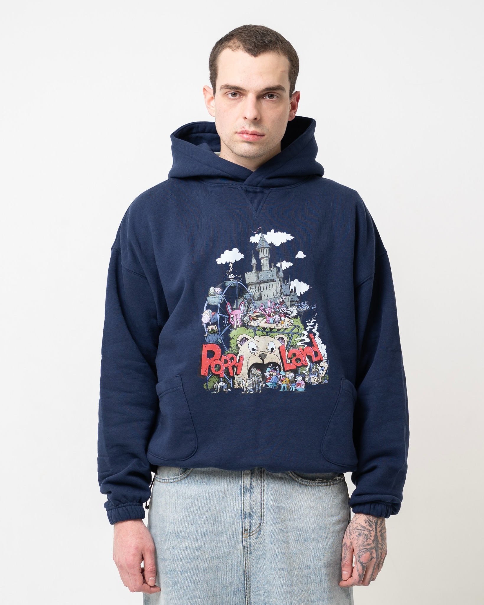 Mężczyzna prezentuje granatową bluzę z kapturem i nadrukiem 'Poppy Land', w dżinsach na białym tle. Widoczny tatuaż na dłoni.