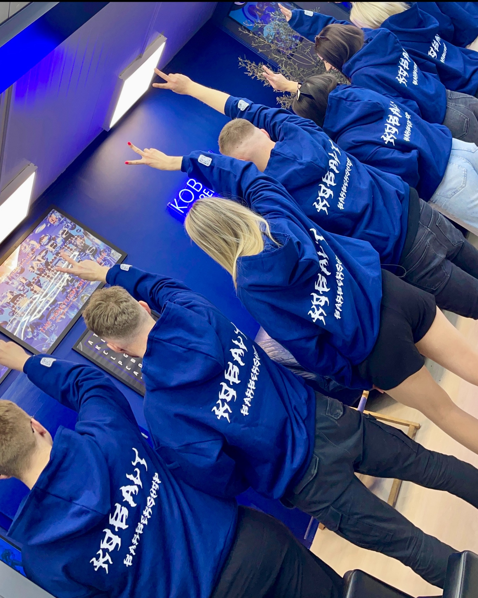 Grupa osób w identycznych, kobaltowych bluzach z kapturem i białym nadrukiem 'KOBALT BARBERSHOP' na plecach, pozujących w niebieskim wnętrzu.