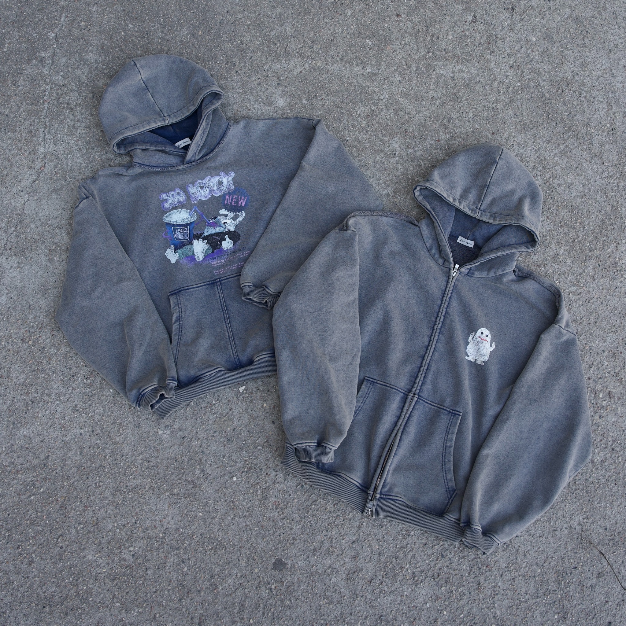 Dwie szare bluzy z kapturem leżące na szarym tle. Jedna bluza rozpinana z małym nadrukiem, druga z dużym nadrukiem graficznym. Bluzy z efektem sprania.