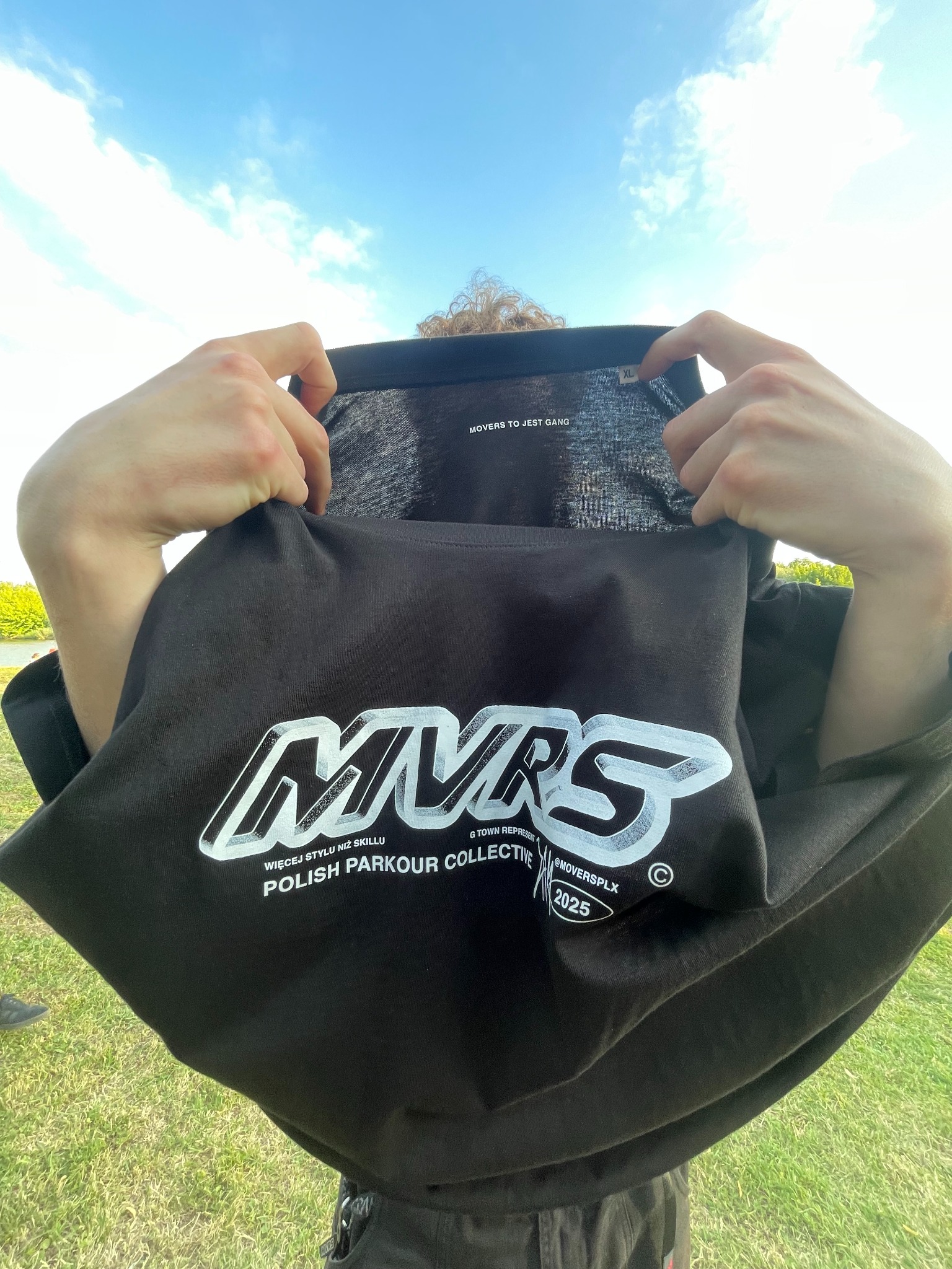 Czarna koszulka z białym nadrukiem 'MVRS' i hasłem 'Polish Parkour Collective', trzymana przez osobę na tle błękitnego nieba i zieleni. Widoczny rozmiar XL.