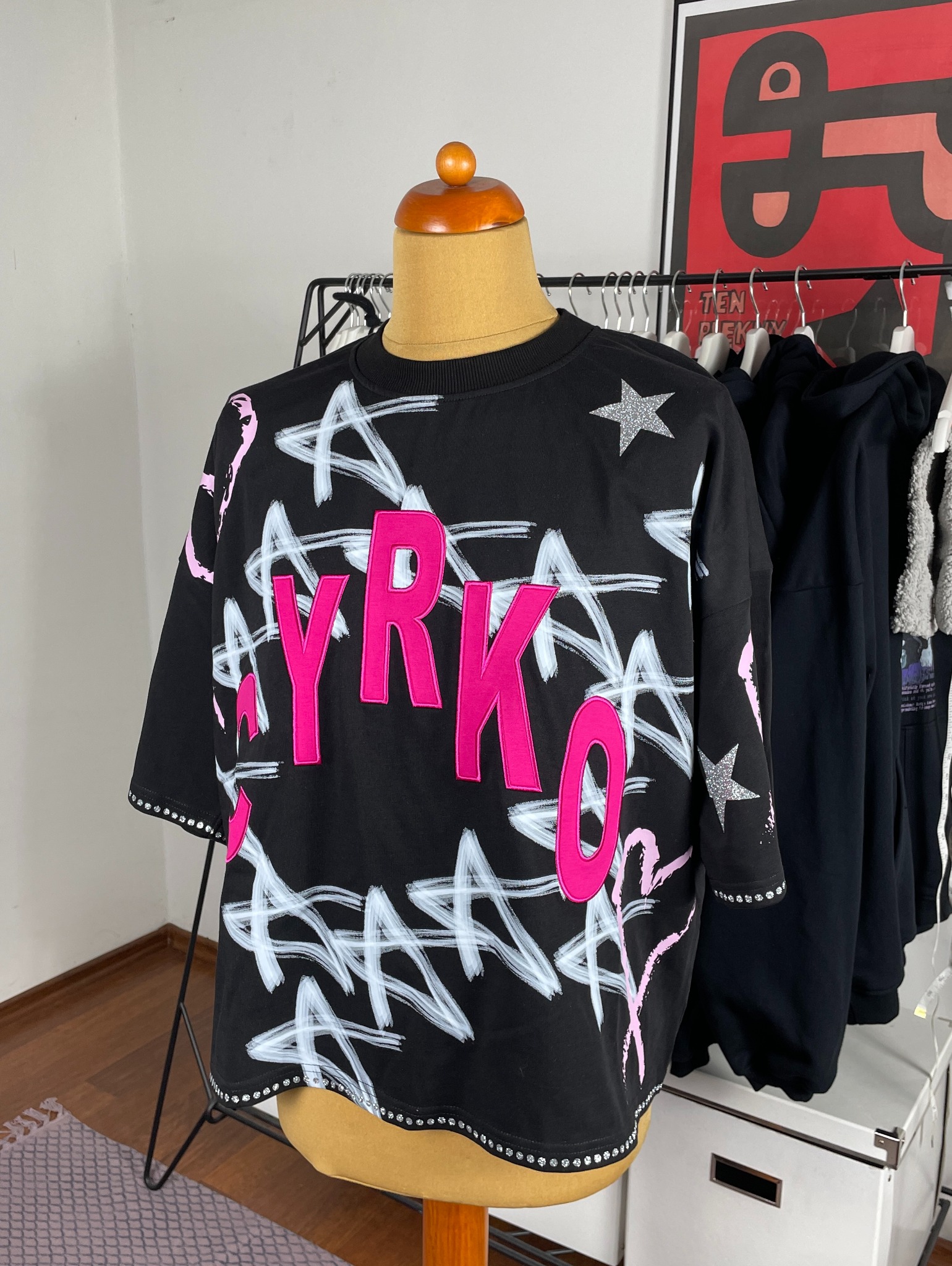 Czarna koszulka z nadrukiem 'CYRKO' w kolorze fuksji, białymi gwiazdami i srebrnymi brokatowymi akcentami na manekinie. Dżety na dole i na rękawach.