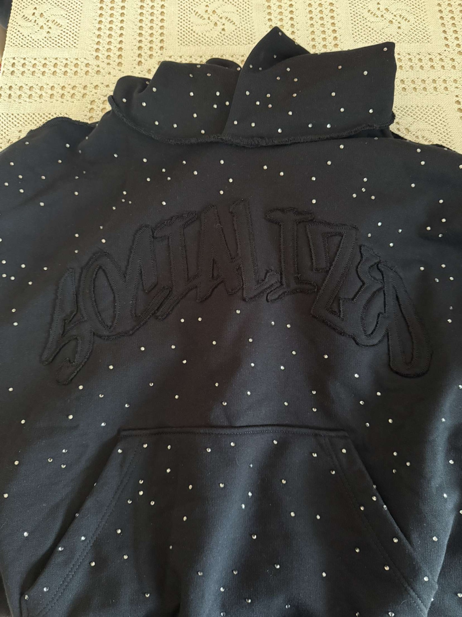 Czarna bluza z kapturem marki odzieżowej, zdobiona haftowanym napisem i licznymi, drobnymi dżetami. Widoczna kieszeń typu kangur i wykończenie dekoltu.