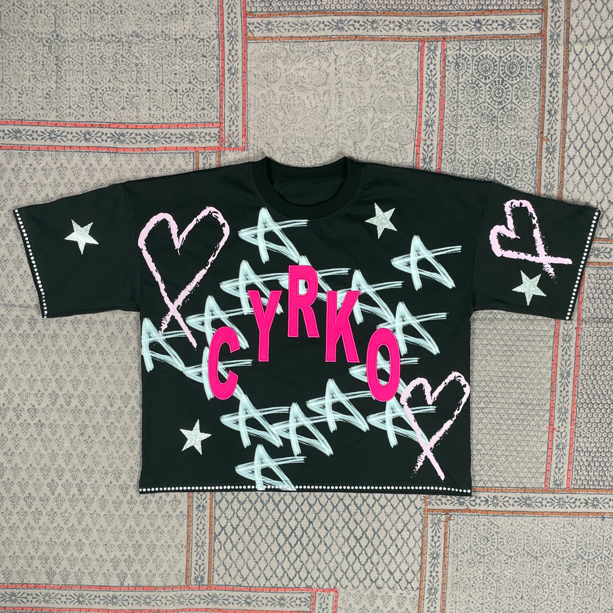 Czarna koszulka z nadrukiem 'CYRKO' w różowych literach, białymi gwiazdami i sercami, leżąca na wzorzystym dywanie. Detale: brokatowe gwiazdy, białe kropki na rękawach.