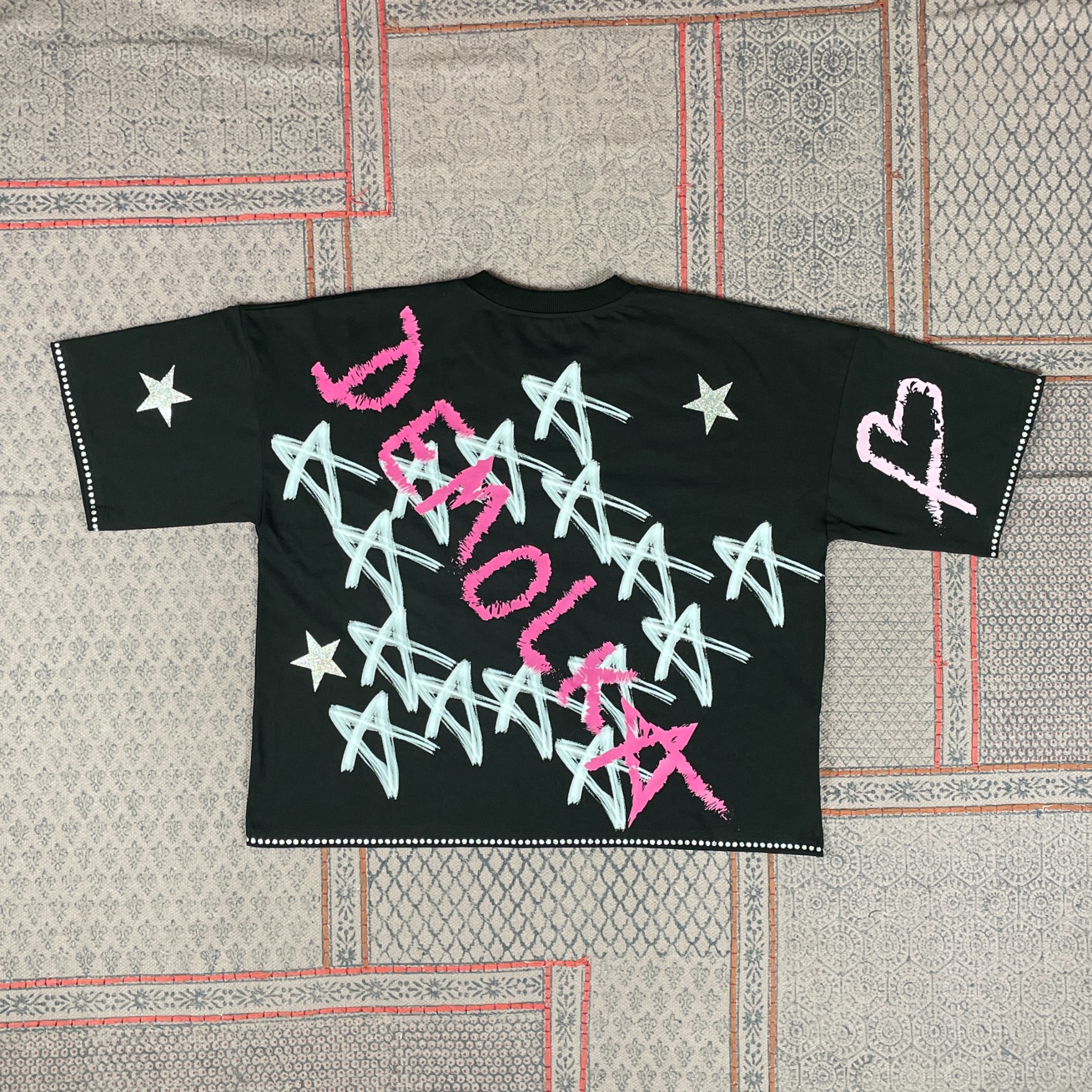 Czarna koszulka z unikatowym nadrukiem gwiazd i napisem w kolorze różowym i białym, leżąca na wzorzystym materiale. Detal: białe kropki na rękawach i dole koszulki.
