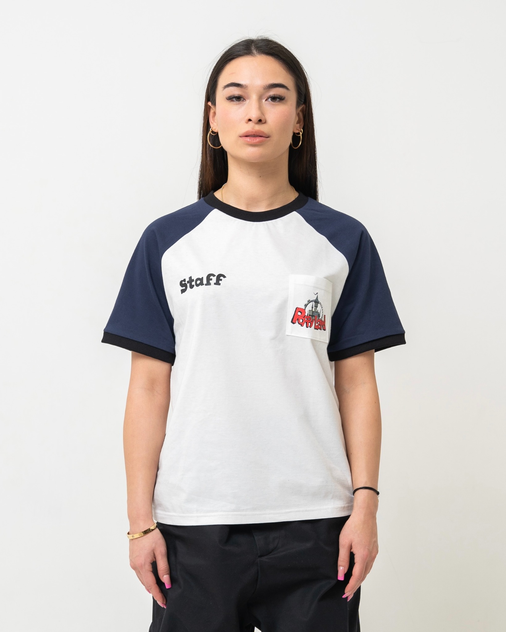 Modelka prezentuje białą koszulkę z granatowymi rękawami raglanowymi, czarnym wykończeniem i nadrukiem 'Staff' oraz kieszonką z logo 'Poppyland'.