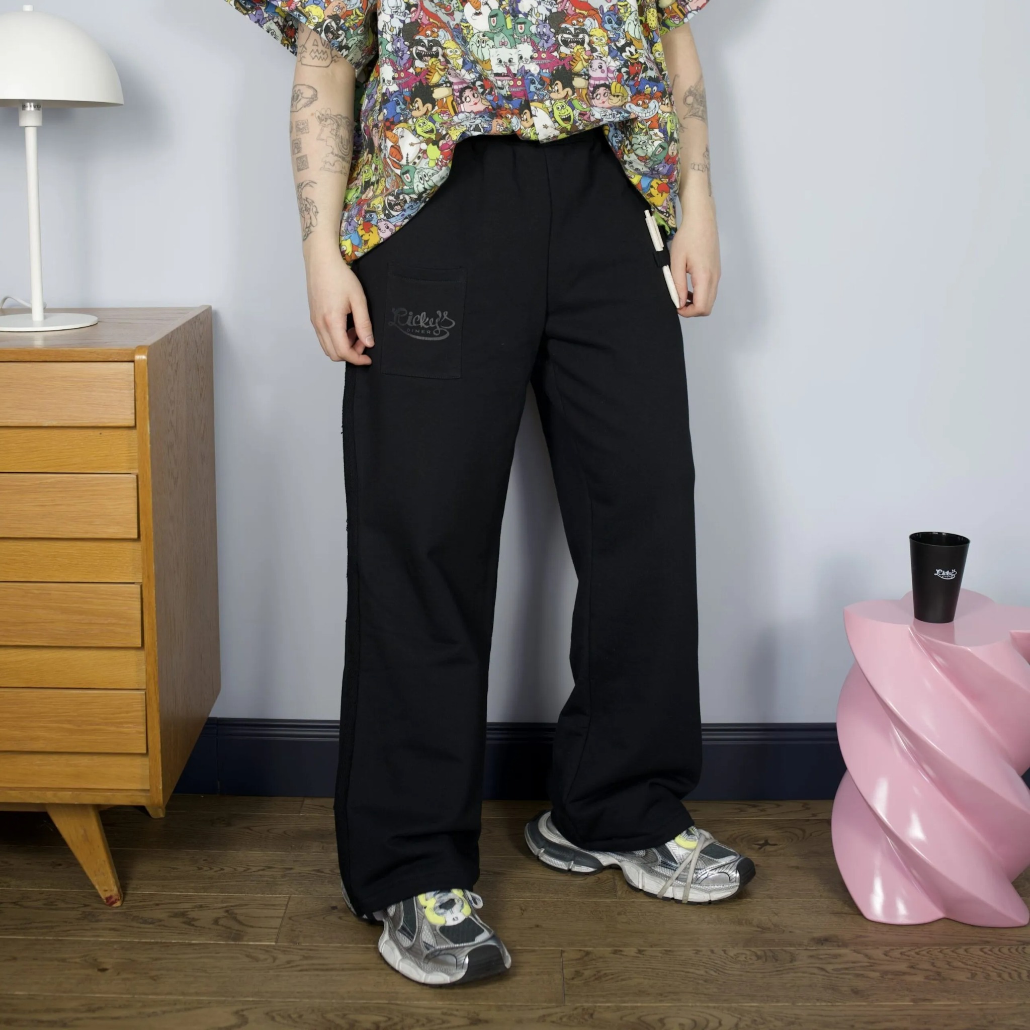 Czarne spodnie dresowe z nadrukiem 'Ricker's Diner' na kieszeni, prezentowane na osobie w kolorowej koszuli, obok różowego stolika i komody.