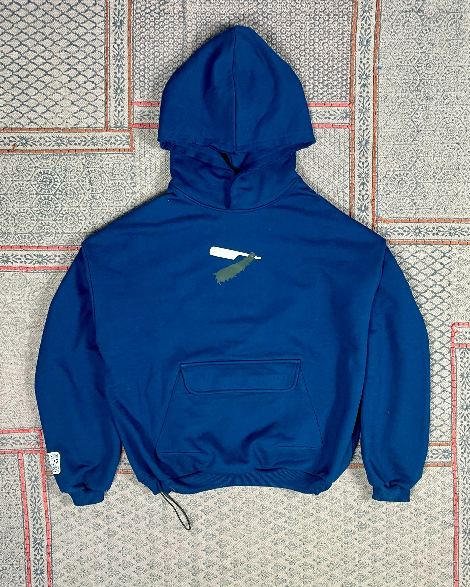 Niebieska bluza z kapturem i nadrukiem brzytwy na tle wzorzystej tkaniny. Widoczna kieszeń na rzep i surowo wykończony kaptur, ściągacz regulowany sznurkiem.