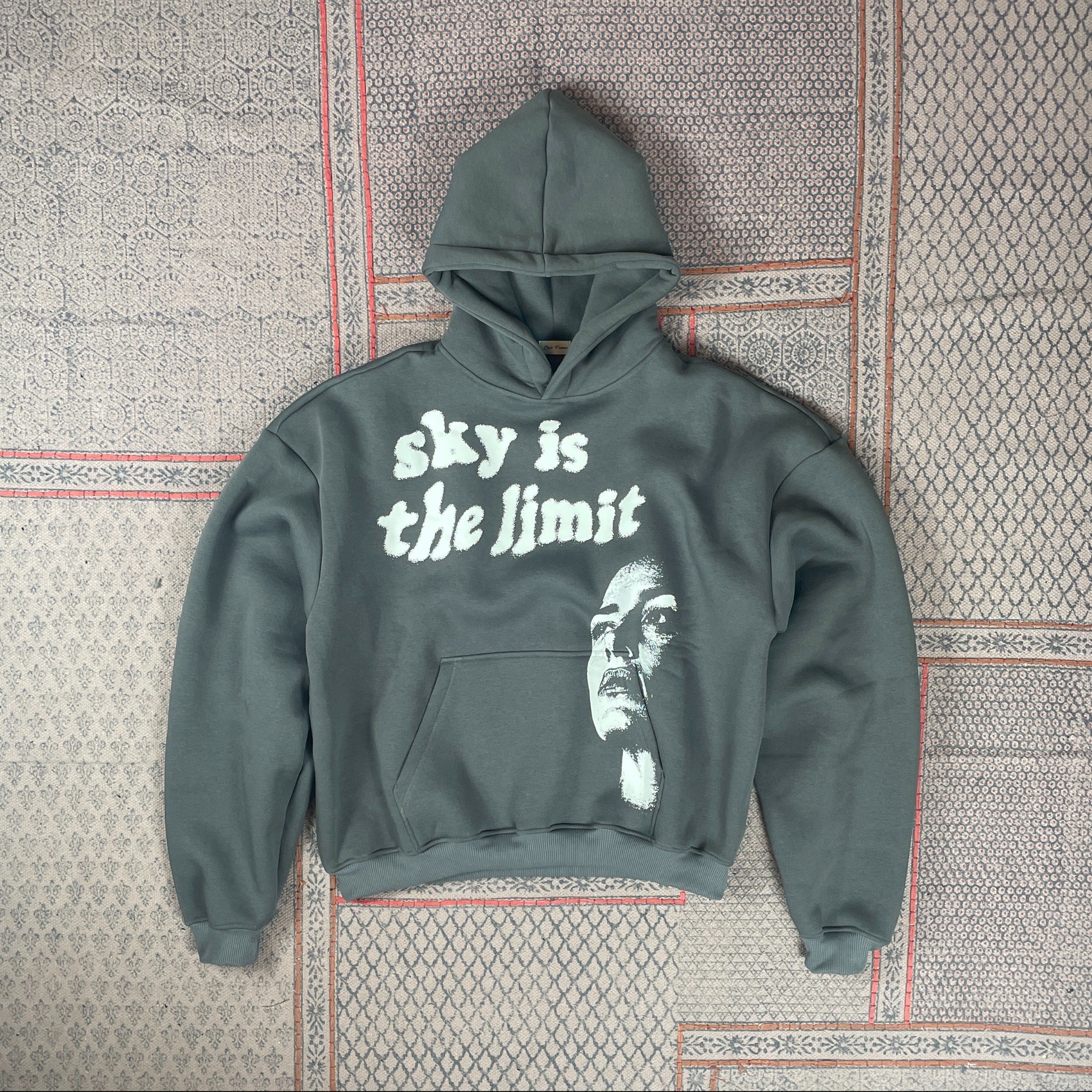 Szaro-zielona bluza z kapturem, z białym nadrukiem 'sky is the limit' i portretem, leży na wzorzystym dywanie. Prezentacja odzieży z nadrukiem.