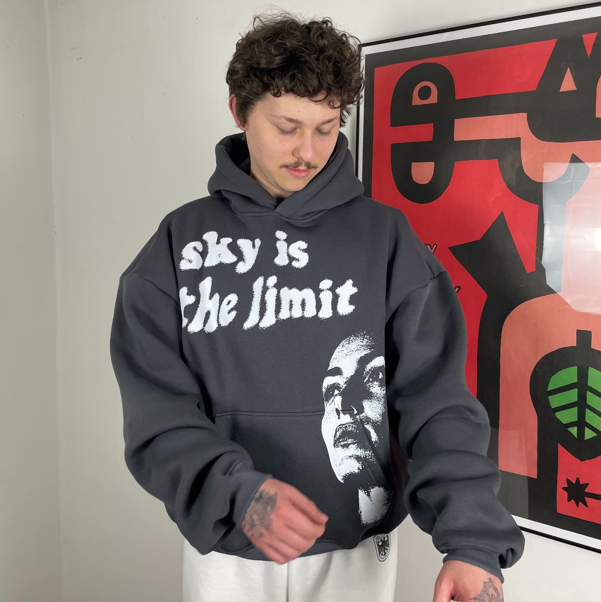 Mężczyzna prezentuje szarą bluzę z kapturem z białym nadrukiem 'sky is the limit' i grafiką twarzy. W tle czerwono-czarny plakat.