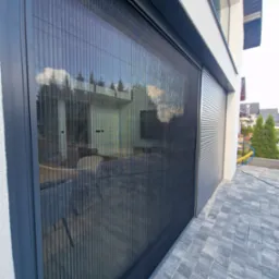 Zewnętrzna, plisowana osłona okienna w nowoczesnym domu. Ciemna rama, szara kostka brukowa, jasna elewacja. Widoczne odbicie otoczenia w osłonie.