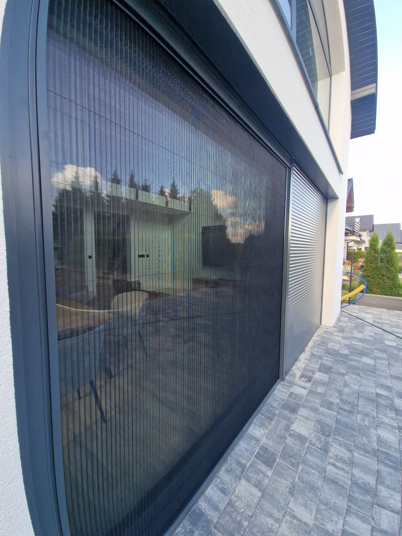 Zewnętrzna, plisowana osłona okienna w nowoczesnym domu. Ciemna rama, szara kostka brukowa, jasna elewacja. Widoczne odbicie otoczenia w osłonie.