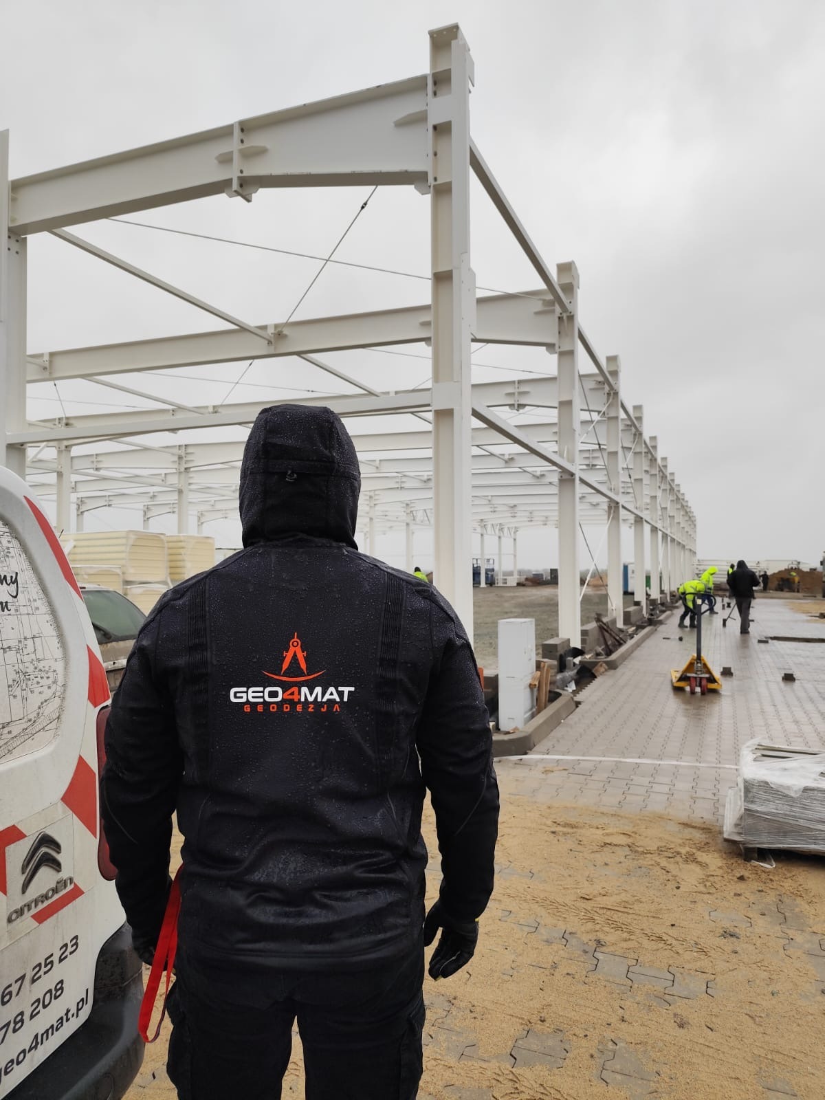 Geodeta w kurtce firmowej GEO4MAT obserwuje montaż konstrukcji stalowej hali. W tle ekipa w żółtych kamizelkach i biała furgonetka Citroen.