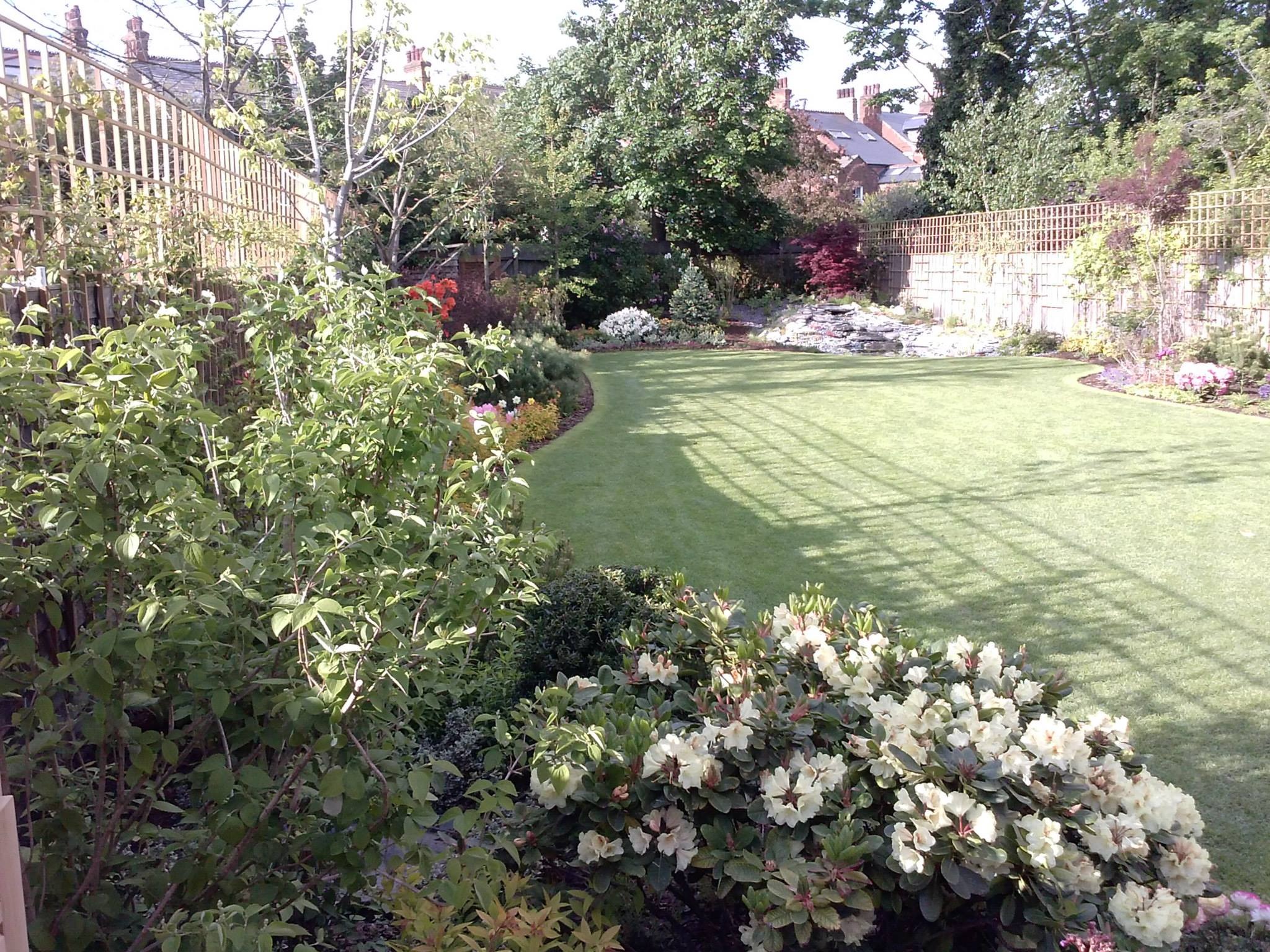 Zadbany ogród z równo przystrzyżonym trawnikiem, rabatami kwiatowymi i ozdobnymi krzewami. W tle widoczne domy z czerwonej cegły i drzewa.