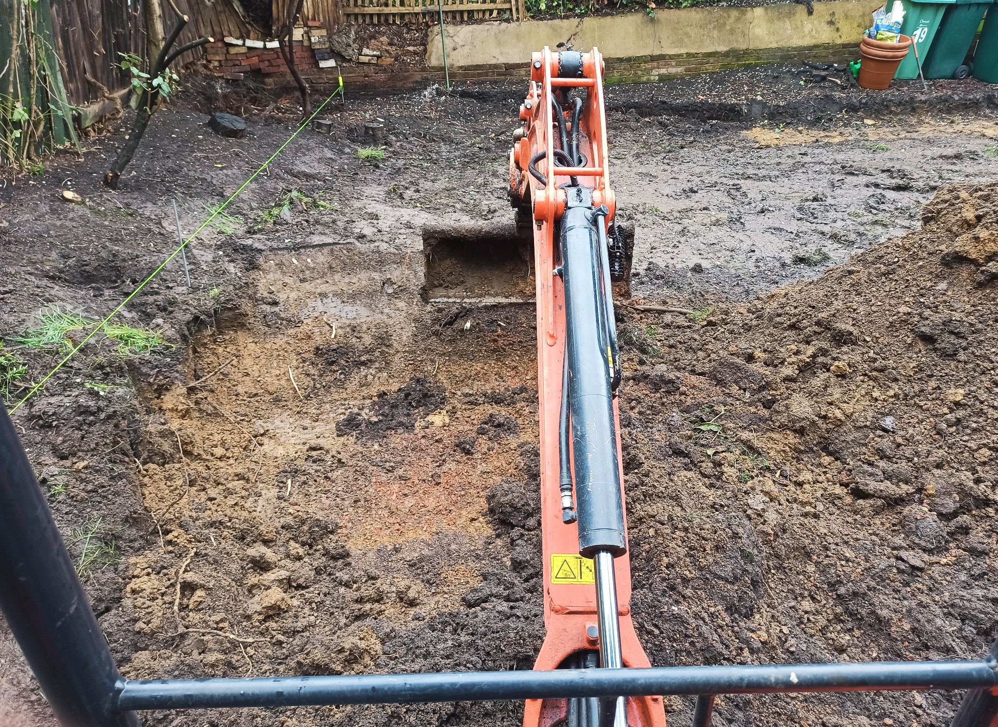 Pomarańczowa mini koparka w trakcie prac ziemnych w ogrodzie. Wykopany dół, widoczna sterta ziemi i wyznaczone granice terenu. Prace przygotowawcze do założenia ogrodu.