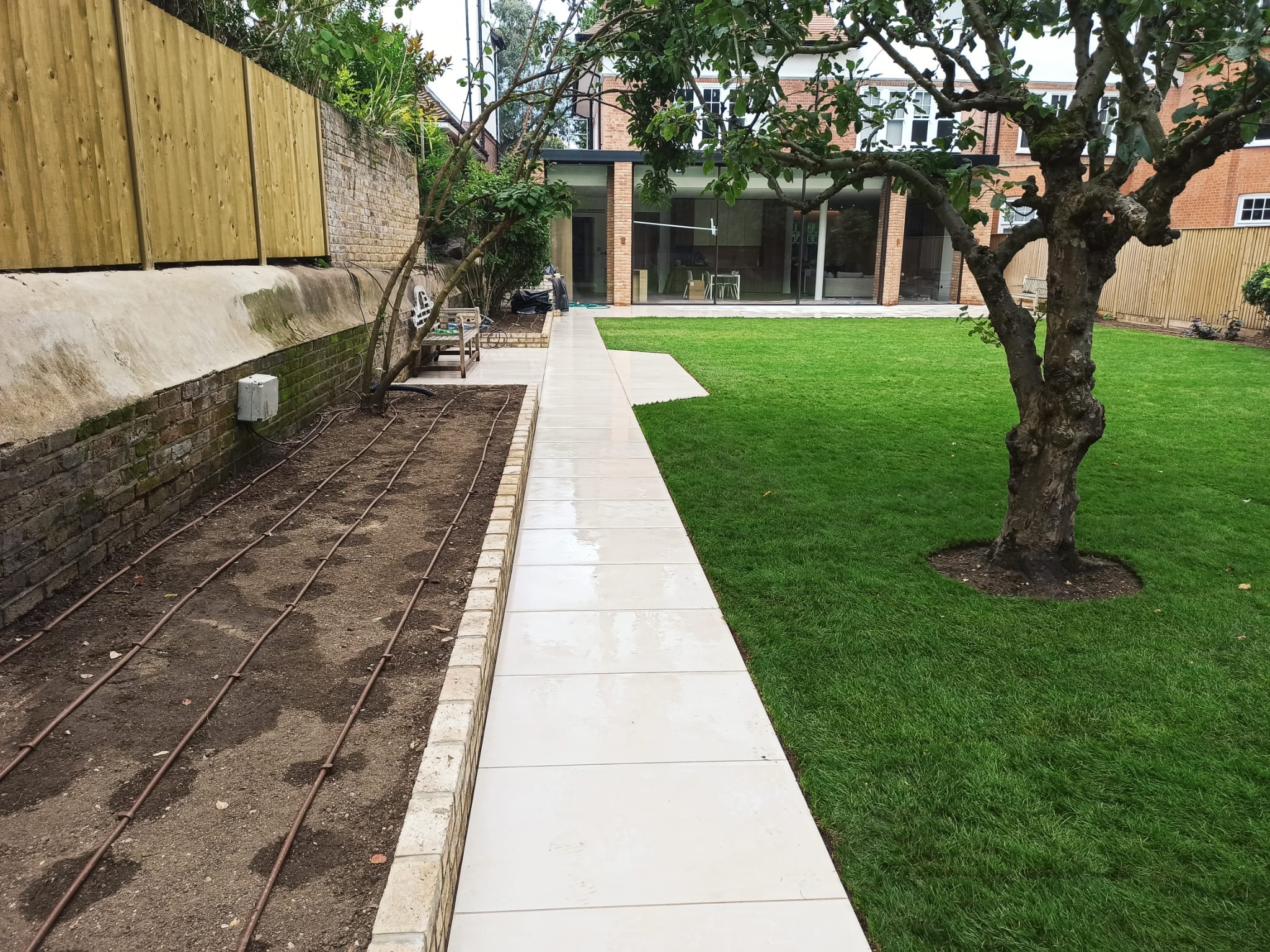 Wyłożony jasnymi płytami chodnik prowadzi przez zadbany ogród z zielonym trawnikiem i przygotowanym miejscem pod nasadzenia. W tle nowoczesny dom.