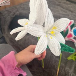 Misja Ruch Malgorzata Zekaj - Ręcznie robiony, biały kwiat z drucików kreatywnych z żółtymi pręcikami, trzymany w dłoni na tle szarej kanapy. Dekoracja DIY, rękodzieło.