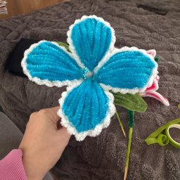 Misja Ruch Malgorzata Zekaj - Ręcznie robiony, duży kwiat z niebieskiego i białego pluszu, trzymany w dłoni na tle szarej narzuty. Widoczne zielone łodygi i liście, oraz fragmenty innych kwiatów.