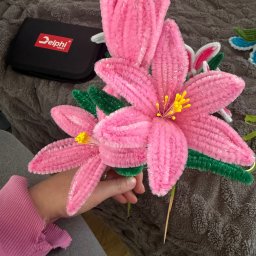 Misja Ruch Malgorzata Zekaj - Ręcznie robione, różowe kwiaty z drucików kreatywnych, trzymane w dłoni, z widocznymi żółtymi pręcikami i zielonymi liśćmi. W tle czarne pudełko Delphi Tools.