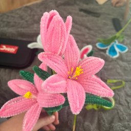 Misja Ruch Malgorzata Zekaj - Ręcznie robione, różowe kwiaty z drucików kreatywnych, trzymane w dłoni, z zielonymi liśćmi i żółtymi pręcikami, na szarym tle.