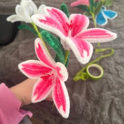 Misja Ruch Malgorzata Zekaj - Ręcznie robione, kolorowe kwiaty z drucików kreatywnych, w odcieniach różu, bieli i zieleni, trzymane w dłoni na tle szarej, teksturowanej powierzchni.