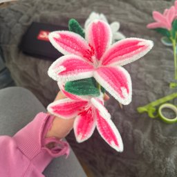 Misja Ruch Malgorzata Zekaj - Ręcznie robione, różowo-białe kwiaty z drucików kreatywnych, trzymane w dłoni na tle szarej kanapy. Widoczne zielone liście i inne kwiaty w tle.