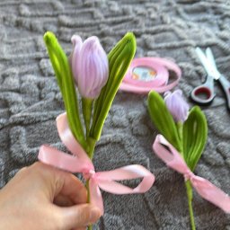 Misja Ruch Malgorzata Zekaj - Ręcznie robione, pluszowe tulipany z zielonymi łodygami i różowymi wstążkami, leżące na szarym kocu. W tle nożyczki i rolka różowej wstążki.