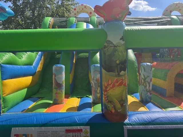 Kolorowy, dmuchany plac zabaw z motywem dinozaurów, zjeżdżalnie i tunele, widok z bliska, w tle zieleń. Idealny na imprezy plenerowe dla dzieci.