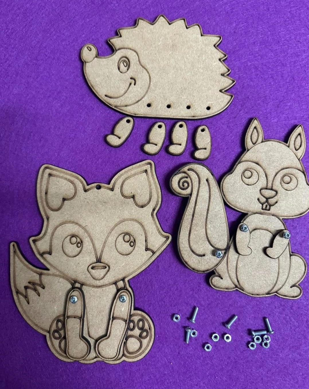 Drewniane, wycięte laserowo elementy do składania figurek zwierząt: jeża, wiewiórki i lisa, wraz ze śrubkami na fioletowym tle. DIY zabawka edukacyjna.